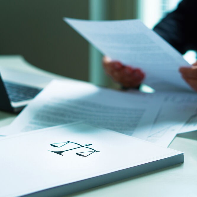 Papierdokumente, auf einem ist eine Waage zur Versinnbildlichung von Gesetz abgebildet, Notebook und Hände, die ein Blatt Papier halten.