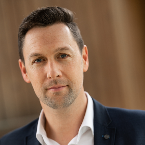 Das Bild zeigt einen Mann in einem formellen Outfit. Er trägt ein weißes Hemd mit Knöpfen, darüber ein marineblaues Sakko. Sein Haar ist dunkel und kurz geschnitten, und er hat blaue Augen.