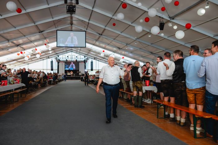 Das Bild zeigt das Innere eines großen Veranstaltungszelts. In der Mitte verläuft ein breiter Gang, der zu einer Bühne am hinteren Ende führt. Links und rechts des Gangs stehen lange, weiß gedeckte Tische, an denen zahlreiche Personen sitzen. Über der Bühne hängt eine große Leinwand, auf der eine Person beim Sprechen zu sehen ist – vermutlich ein Live-Übertragungsbild. Die Decke des Zelts ist festlich mit weißen und roten Luftballons geschmückt. Ein Mann geht den Mittelgang entlang, während auf der rechten Seite mehrere Personen stehen und applaudieren. Die Atmosphäre wirkt feierlich und gemeinschaftlich.
