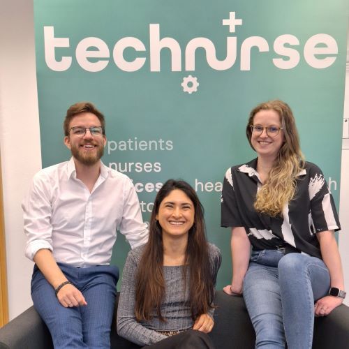 Drei Personen sitzen vor einer Wand mit dem Schriftzug 'technurse' und weiteren englischen Textzeilen