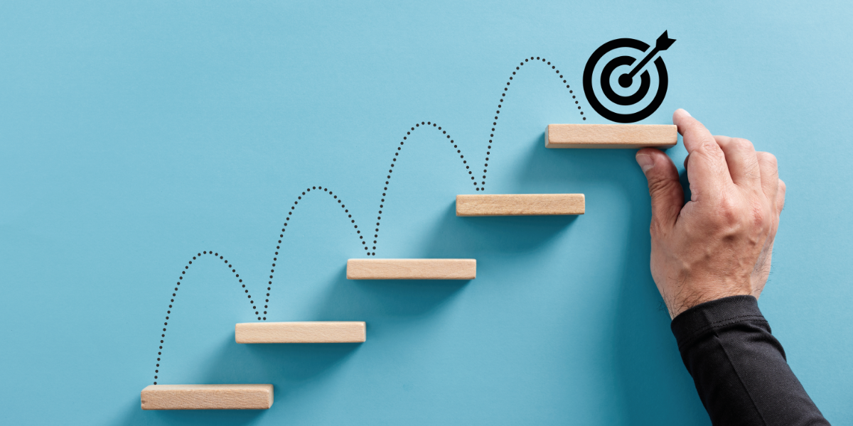 Hand platziert Holzstufen nach oben, eine gepunktete Linie führt zu einer Zielscheibe – Symbol für Fortschritt und Zielerreichung.