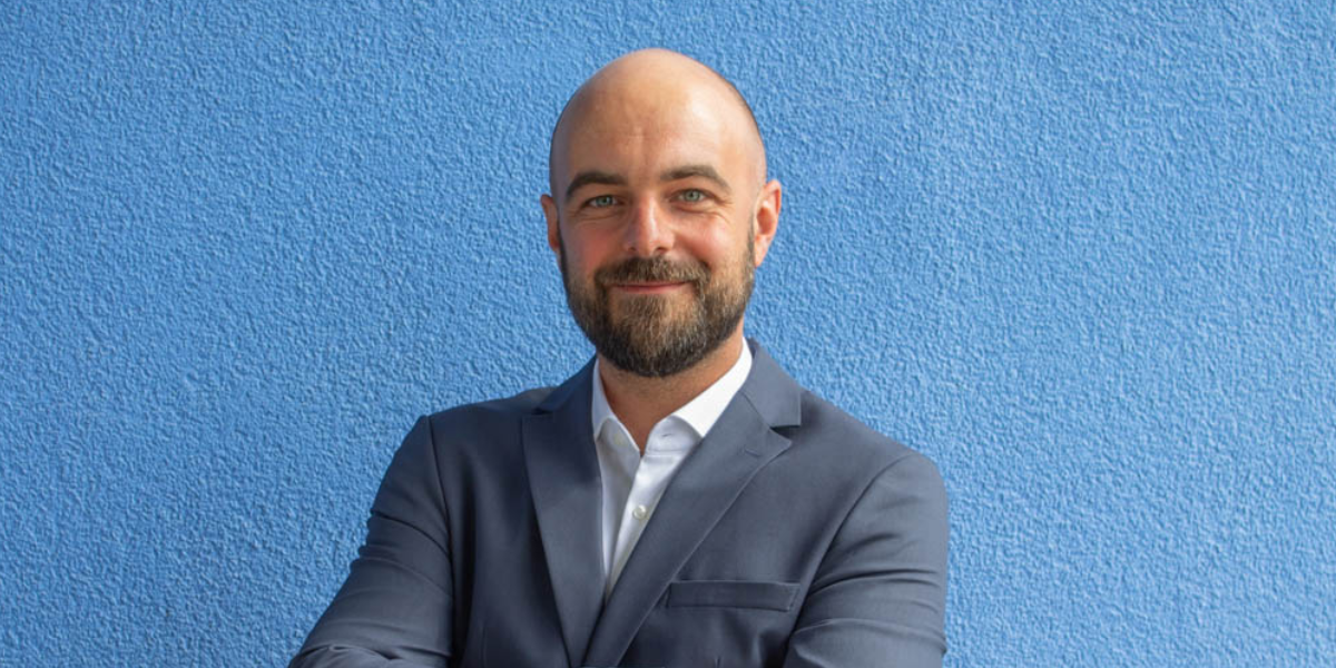 Ergebnis der Bildbeschreibung Das Bild zeigt einen Mann mit Bart und Anzug vor einer blauen Wand. Er lächelt in die Kamera.