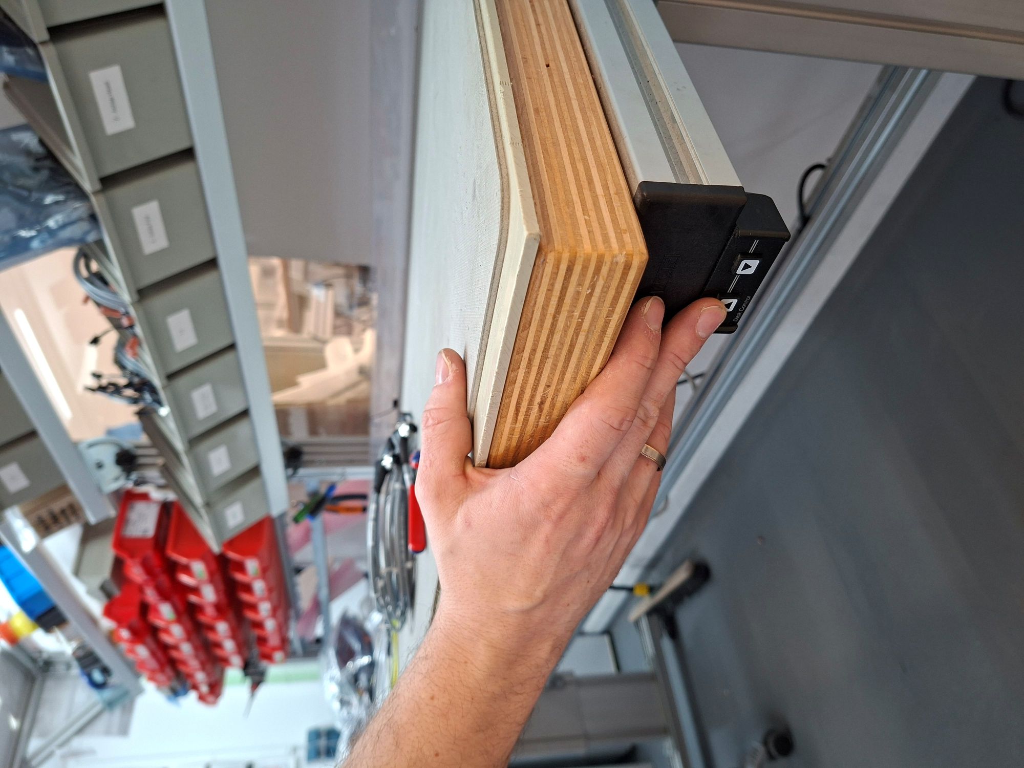 Hand greift an die Kante eines höhenverstellbaren Arbeitstisches mit Holzplatte und Metallgestell in einer Werkstatt