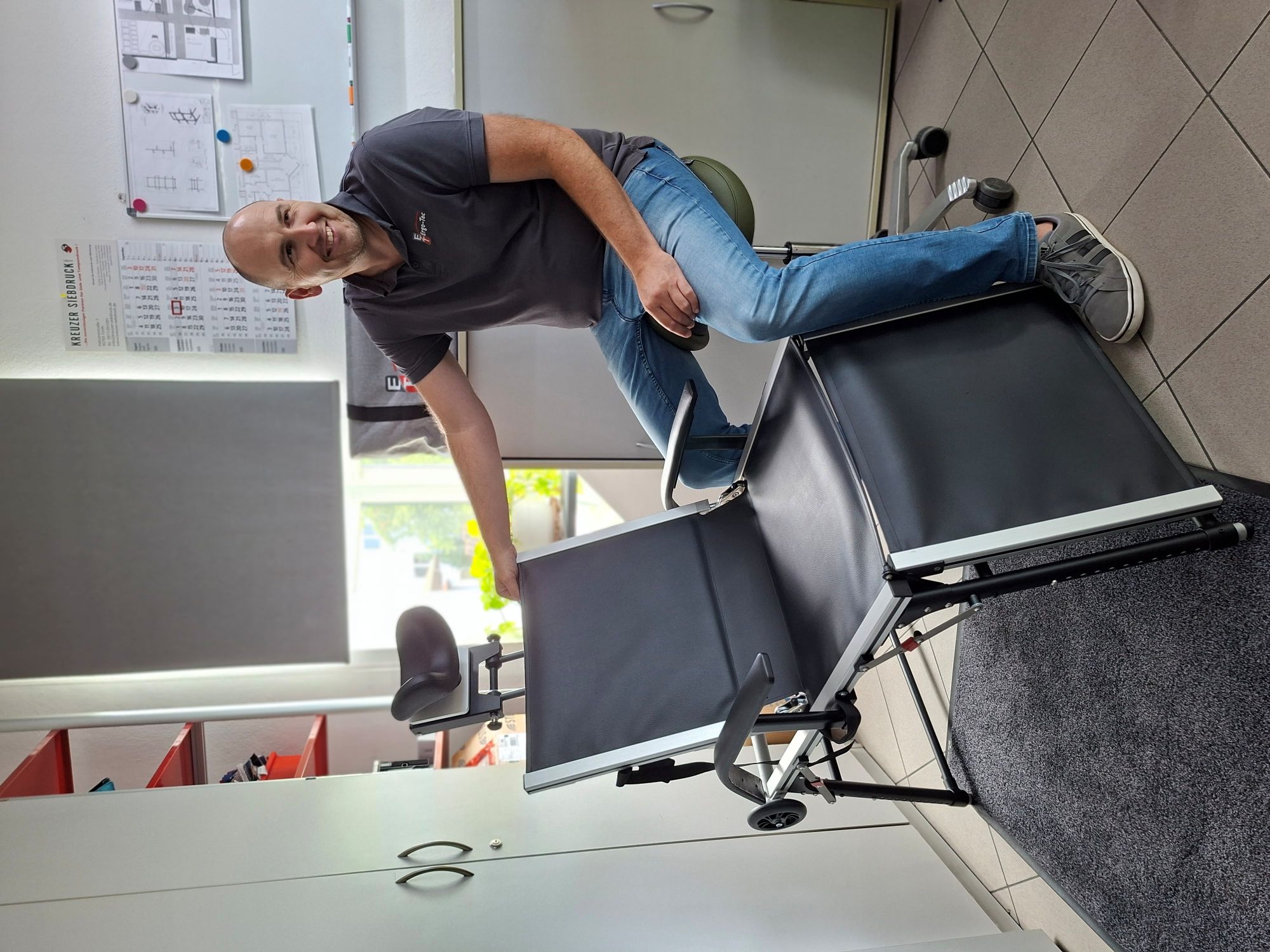 Mann sitzt auf einem Bürostuhl neben einem schwarzen, verstellbaren Therapiesessel in einem hellen Raum mit Schränken und Kalender an der Wand