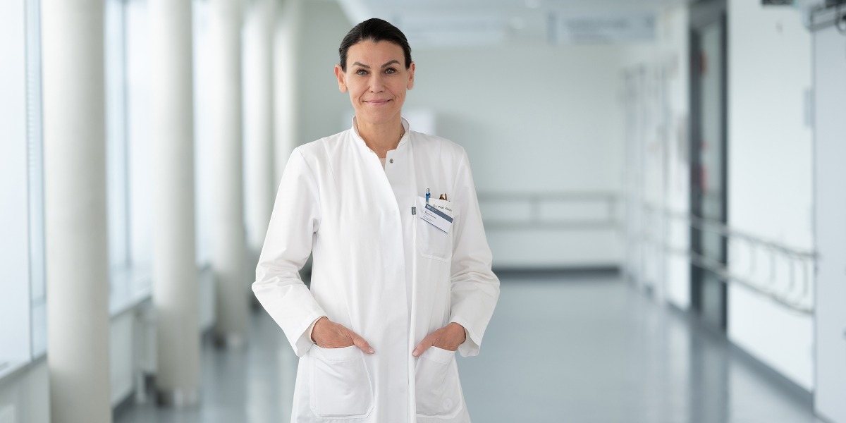 Person in weißem Arztkittel mit Namensschild und Stiften in den Brusttaschen steht in einem hellen Flur mit großen Fenstern