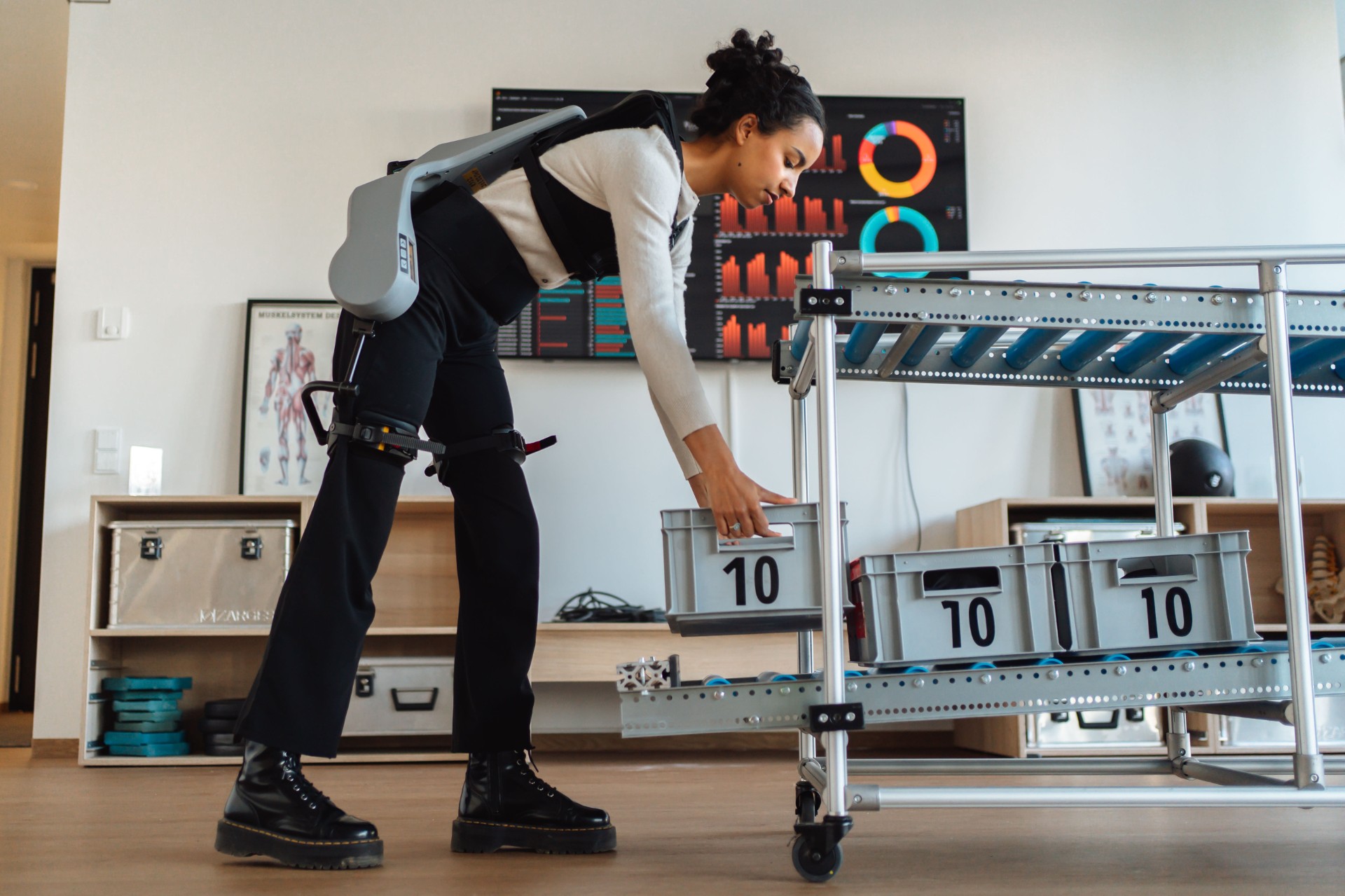 Person mit einem Exoskelett an den Beinen greift nach einer Kiste mit der Aufschrift '10' in einem Raum mit Regalen und medizinischen Postern an der Wand