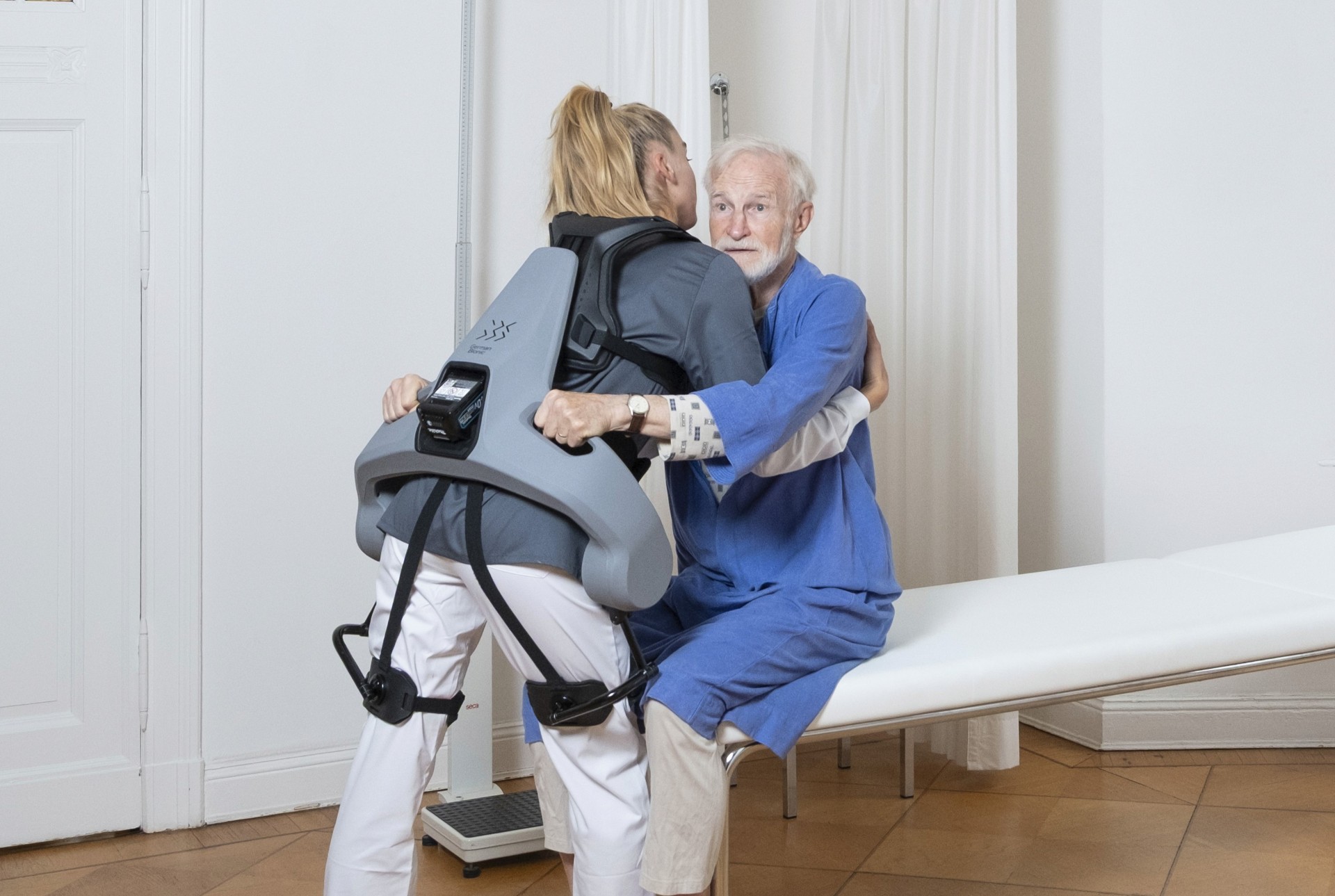 Person mit Exoskelett hebt eine andere Person vom Stuhl in einem Raum mit Holzboden und weißen Wänden