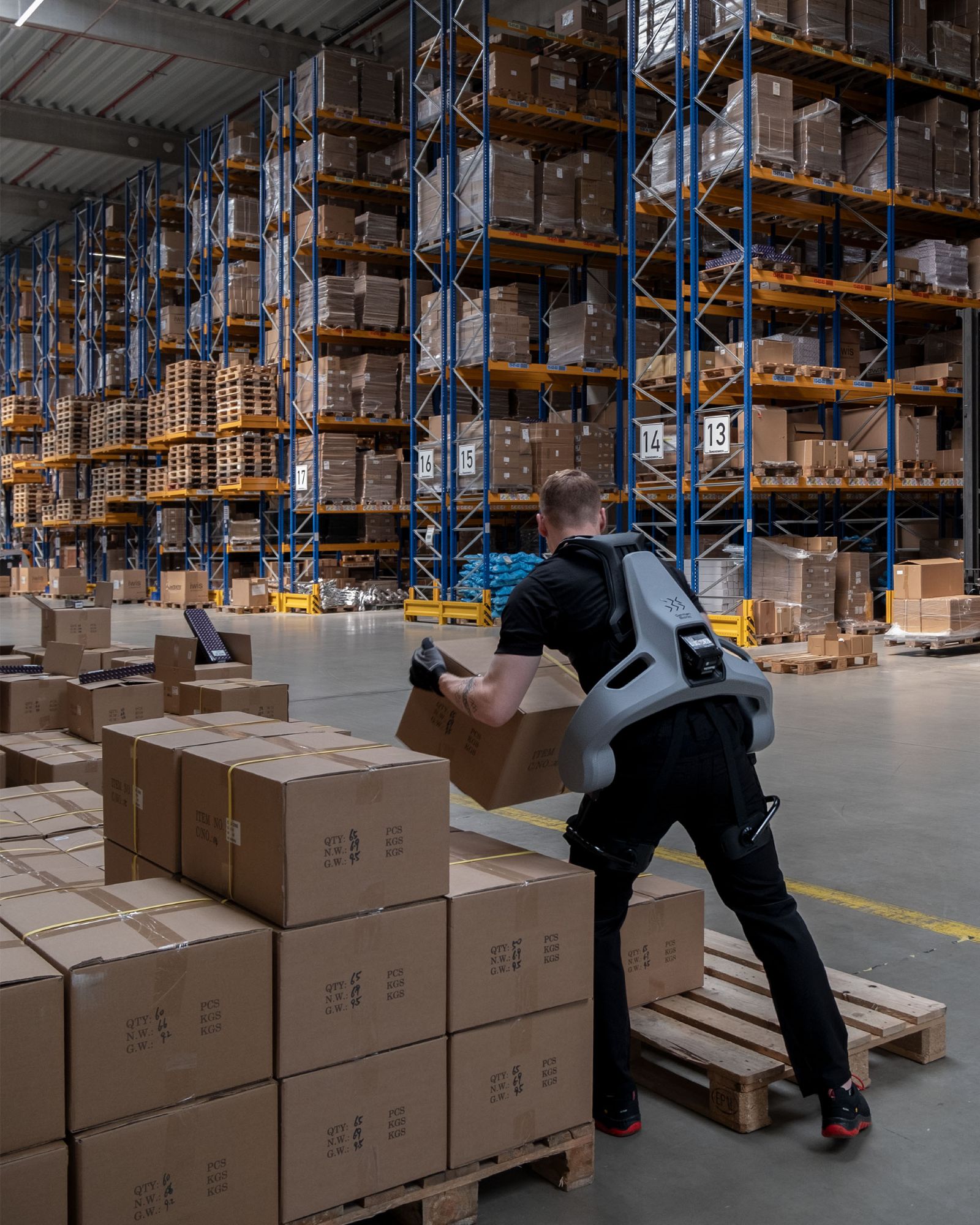 Mann mit Exoskelett hebt Karton in großem Lager mit hohen Regalen und vielen Kartons