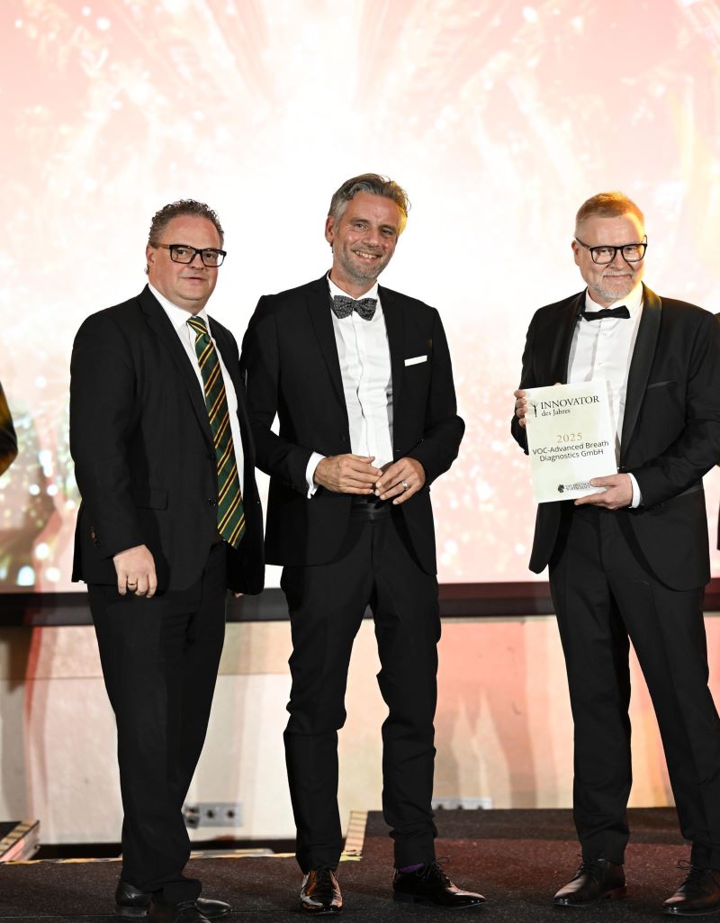 Drei Personen in eleganten Anzügen stehen auf einer Bühne. Eine Person hält ein gerahmtes Zertifikat mit der Aufschrift „INNOVATOR DES JAHRES“ und weiteren Details. Der Hintergrund zeigt ein leuchtendes, festliches Muster in Gold- und Orangetönen.