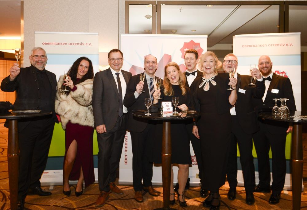 Eine Gruppe von Personen steht um zwei Stehtische in einem Empfangsbereich. Alle tragen formelle Kleidung und halten Gläser mit Sekt für einen Toast. Im Hintergrund sind zwei Banner mit der Aufschrift „Oberfranken offensiv e.V.“ sowie ein Logo sichtbar.