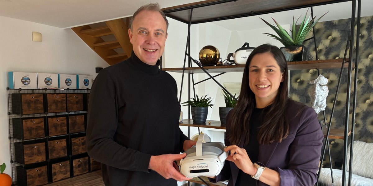 Das Bild zeigt zwei Personen, einen Mann und eine Frau, die lächeln und eine VR-Brille mit der Aufschrift "magic horizons" halten. Sie stehen vor einem Regal mit verschiedenen dekorativen Objekten wie Pflanzen, einer goldenen Kugel und einer Art Skulptur. Im Hintergrund befinden sich weitere Regale mit Kisten.