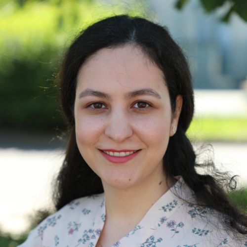 Person mit langen, dunklen, welligen Haaren trägt ein helles, geblümtes Oberteil und steht vor unscharfem, grünem Hintergrund im Freien