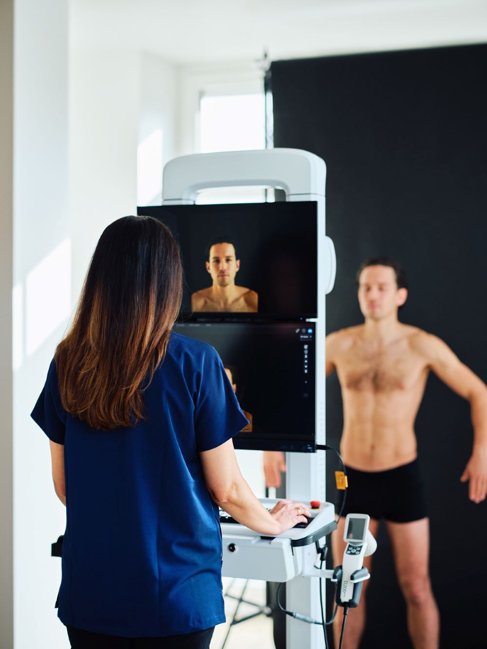 Person steht vor einem medizinischen Scanner, Fachkraft bedient das Gerät in einem hellen Raum.