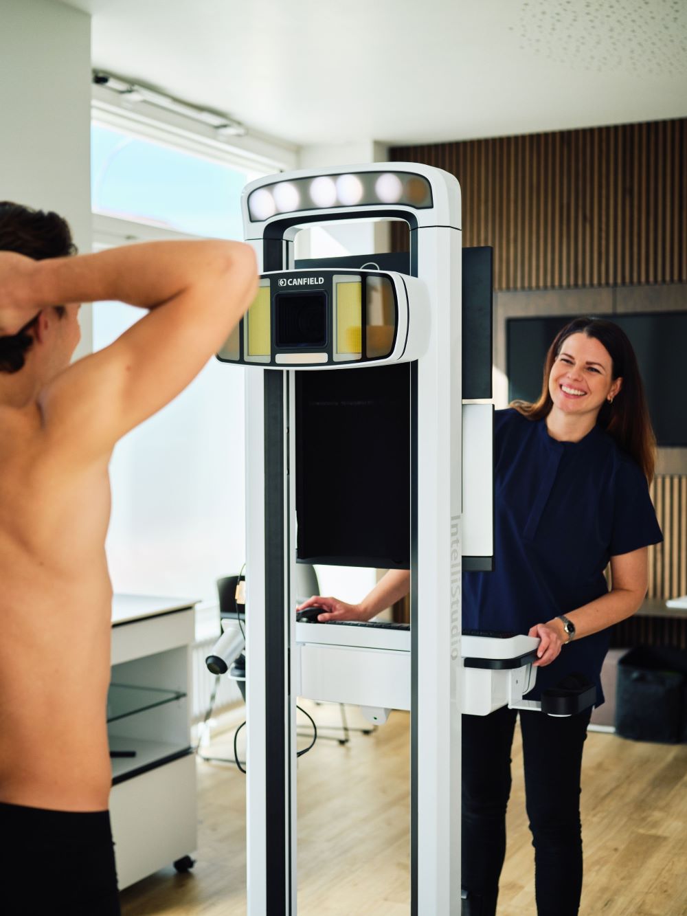 Medizinische Untersuchungsszene mit einer stehenden Person in Unterwäsche vor einem schwarzen Hintergrund und einer Fachkraft an einem Gerät.