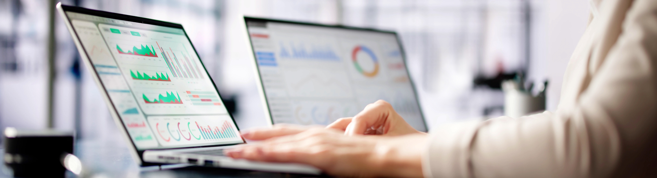 Auf der Grafik sieht man eine Person, die an einem Laptop arbeitet, auf dessen Bildschirm Diagramme und Analysen zur Datenauswertung angezeigt werden; im Hintergrund steht ein weiterer Monitor mit grafischen Auswertungen.