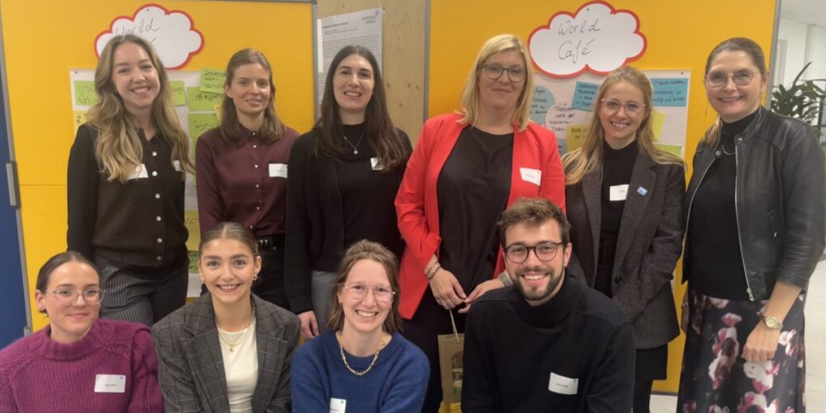 Das Bild zeigt eine Gruppe von elf Personen, die in zwei Reihen vor einer gelben Wand posieren. Auf der Wand sind Notizen und eine Sprechblase mit der Aufschrift "World Café" angebracht.