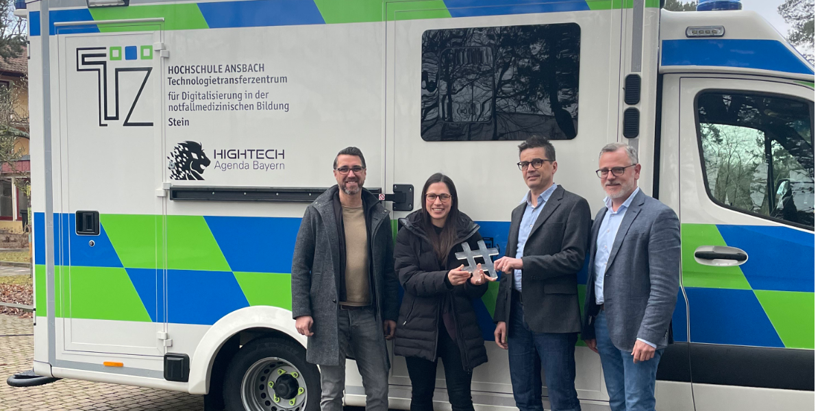 Vier Personen stehen vor einem weißen Fahrzeug mit blau-grünen Streifen und dem Schriftzug "HOCHSCHULE ANSBACH TechnologieTransferZentrum für Digitalisierung in der notfallmedizinischen Bildung Stein"