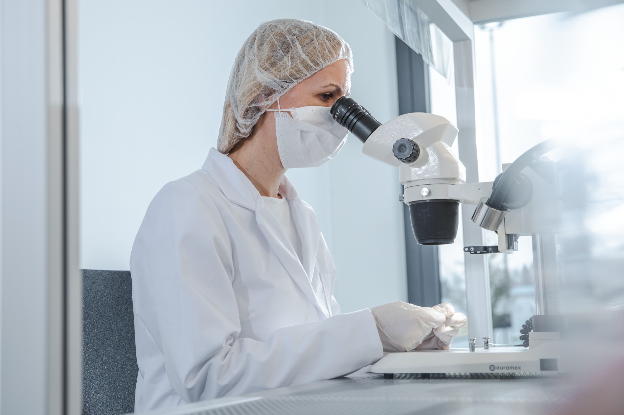 Person mit Haarnetz und Schutzbrille sitzt an Mikroskop in Laborumgebung