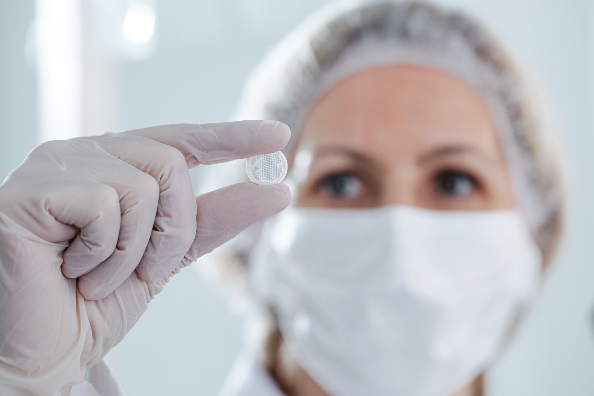 Hand mit Einweghandschuh hält eine kleine transparente Kapsel vor unscharfem Hintergrund