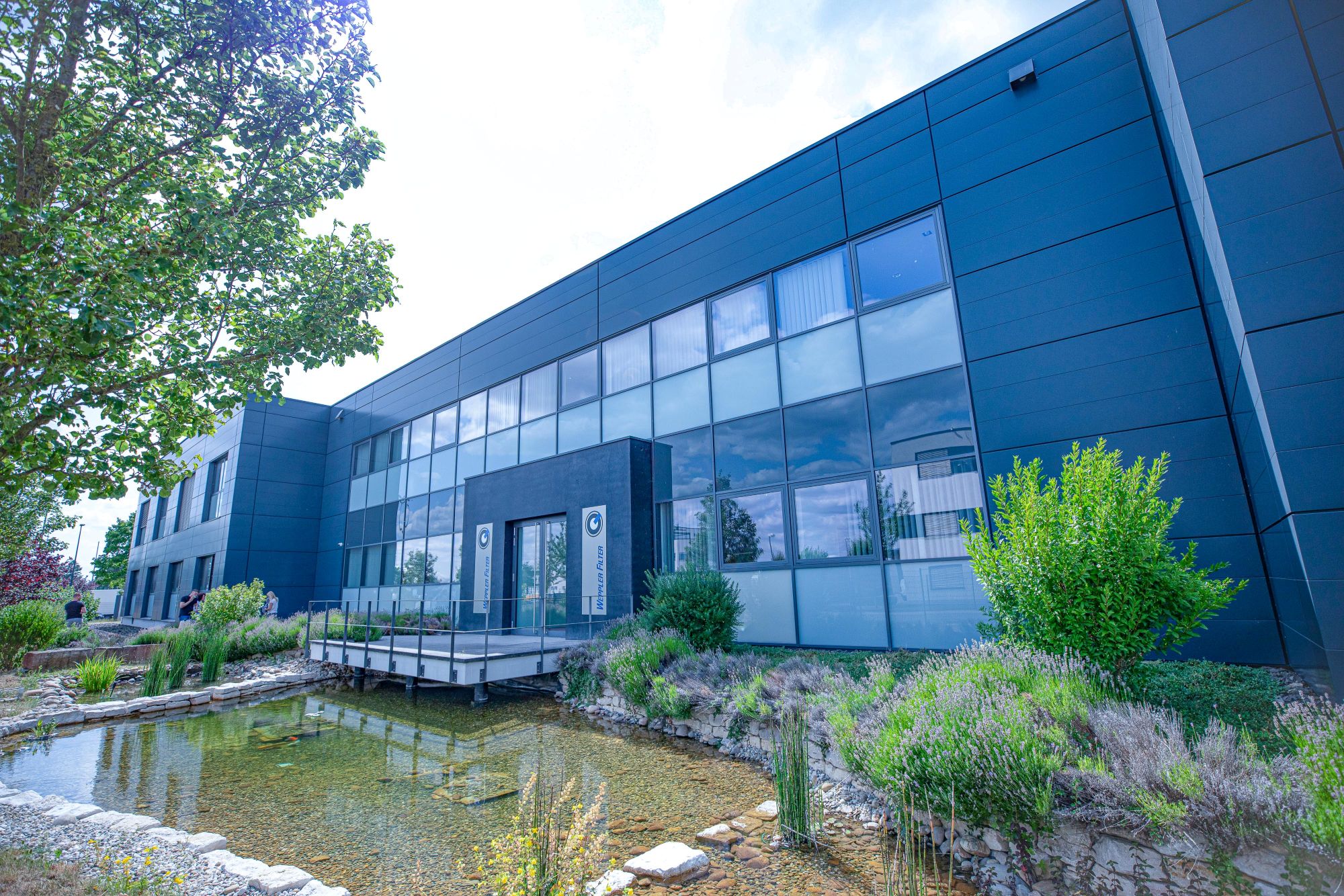 Modernes Firmengebäude mit großer Glasfassade, davor ein Teich und umgebende Bepflanzung