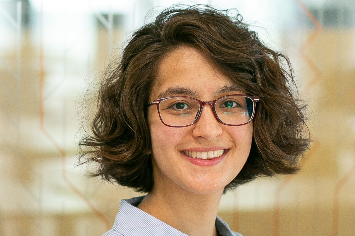 Das Bild zeigt das Porträt einer jungen Frau. Sie hat dunkles, gewelltes Haar und trägt eine Brille mit einem roten Rahmen. Sie lächelt freundlich und zeigt ihre Zähne. Sie trägt ein hellblaues Hemd mit weißen Streifen.