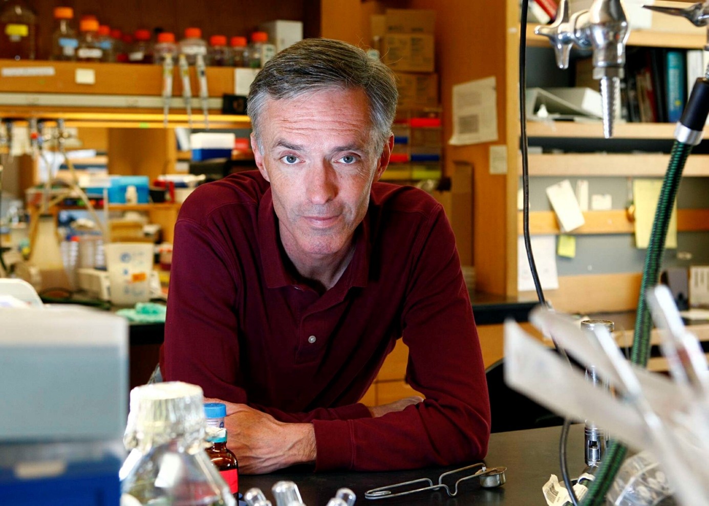 Mann mit grauen Haaren und rotem Pullover lehnt in einem Labor an einem Tisch mit Laborgeräten und Reagenzgläsern