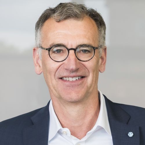 Mann mittleren Alters mit kurzen grauen Haaren, Brille und dunklem Sakko lächelt in die Kamera