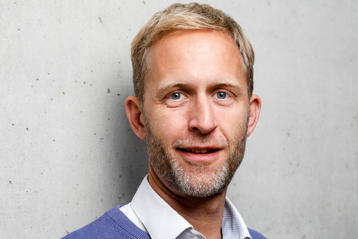 Porträt eines lächelnden Mannes mit kurzem blonden Haar und Bart vor einer hellen Wand