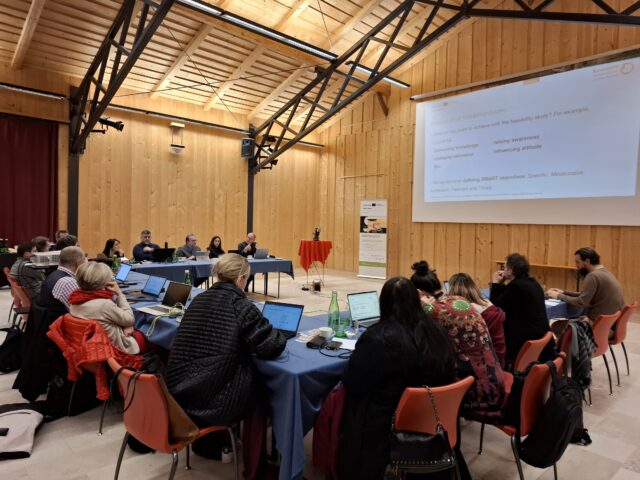 Ein Konferenzraum mit Tischen in U-Form, an denen viele Menschen sitzen und auf eine Powerpoint blicken in einem Holzgebäude. 