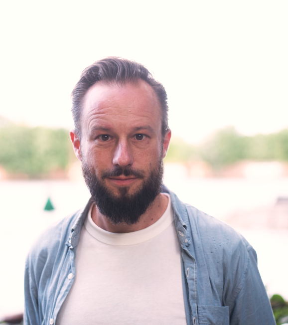 Ein Mann mit weißen T-Shirt, Jeanshemd und Bart schaut frontal in die Kamera.