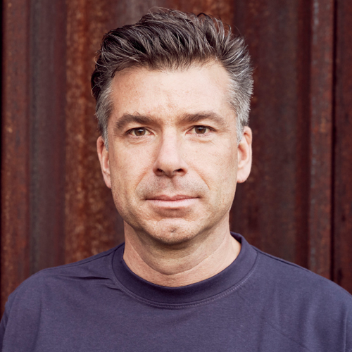 Ein Portraitbild. Ein Mann mit leicht gräulichen Haar blickt frontal in die Kamera. Er trägt einen blauen Pullover und steht vor einer braunen Wand.