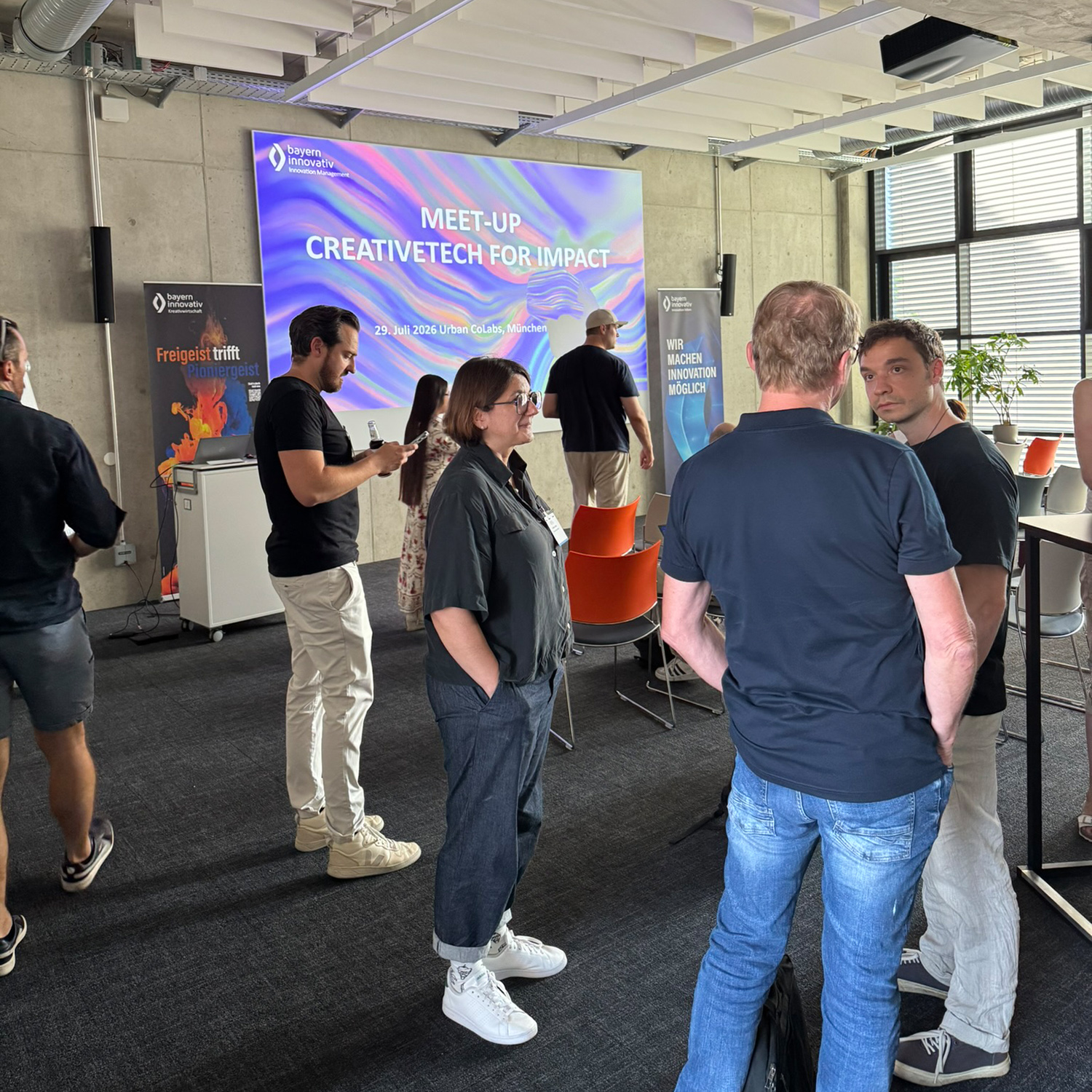 Einige der Teilnehmenden des Meet-ups, darunter Johanna Lison, Projektmanagerin Crea-tiveTechHUB der Bayern Innovativ GmbH, stehen im Raum und unterhalten sich. Im Hin-tergrund sind zwei Roll-ups, die Leinwand und rote Stühle zu sehen.