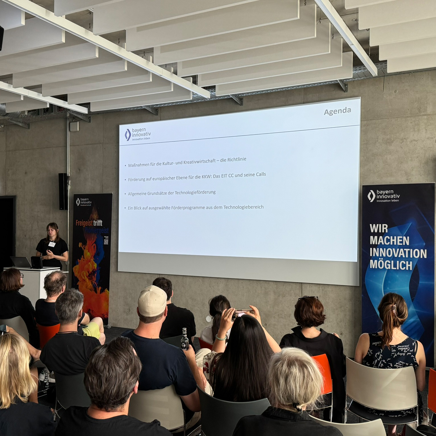Charlotte Stegmayer, Projektmanagerin CreativeTechHUB der Bayern Innovativ GmbH, steht an einem Rednerpult und spricht zu den Teilnehmenden des Meet-ups. Im Hintergrund stehen zwei Roll-ups der Bayern Innovativ GmbH.