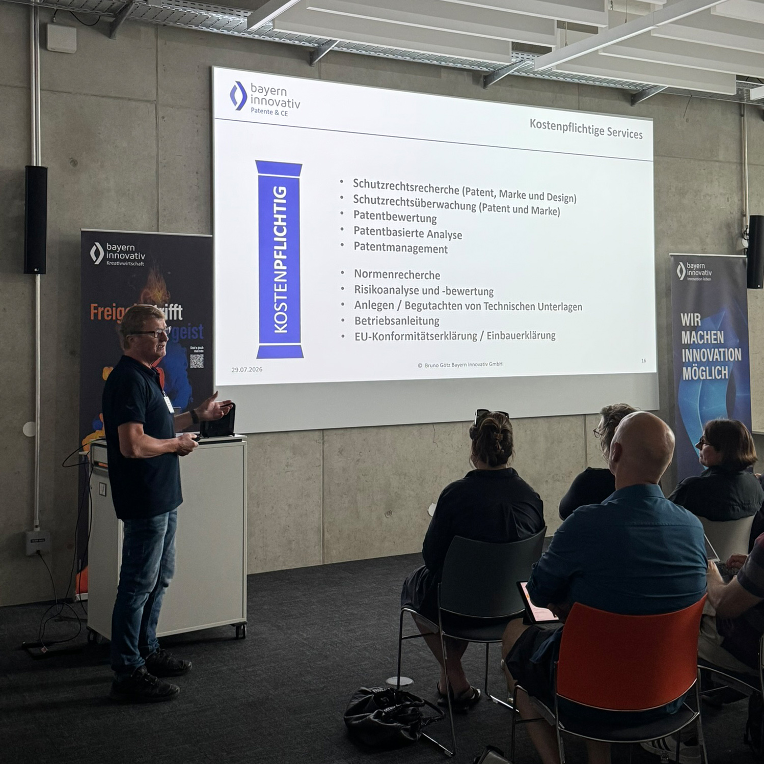 Bruno Götz, Leiter Patente und Normen bei der Bayern Innovativ GmbH, steht neben dem Rednerpult und referiert vor den Teilnehmenden des Meet-ups und zeigt die wichtigsten Schutzrechte für Kultur- und Kreativschaffende auf. Im Hintergrund sind ein Roll-up und eine Leinwand zu sehen.