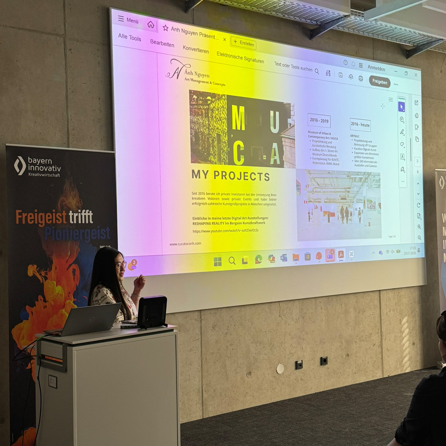 Ánh Nguyen steht neben dem Rednerpult und informiert die Teilnehmenden des Meet-ups über das Thema Curator & Art Consultant. Im Hintergrund sind ein Roll-up und eine Leinwand zu sehen. Die Leinwand wird von mehreren bunten Farben beleuchtet. 