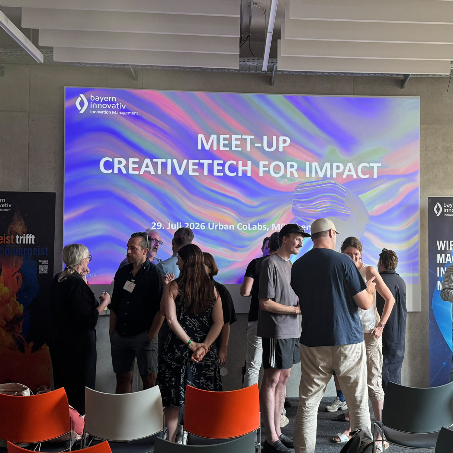 Die Teilnehmenden des Meet-ups stehen vor einer großen Leinwand und unterhalten sich angeregt. Mehrere Stühle in den Farben rot, weiß und grau stehen im Raum. Zwei Roll-ups stehen links und rechts vor der Wand und dazwischen ist die Leinwand angebracht.