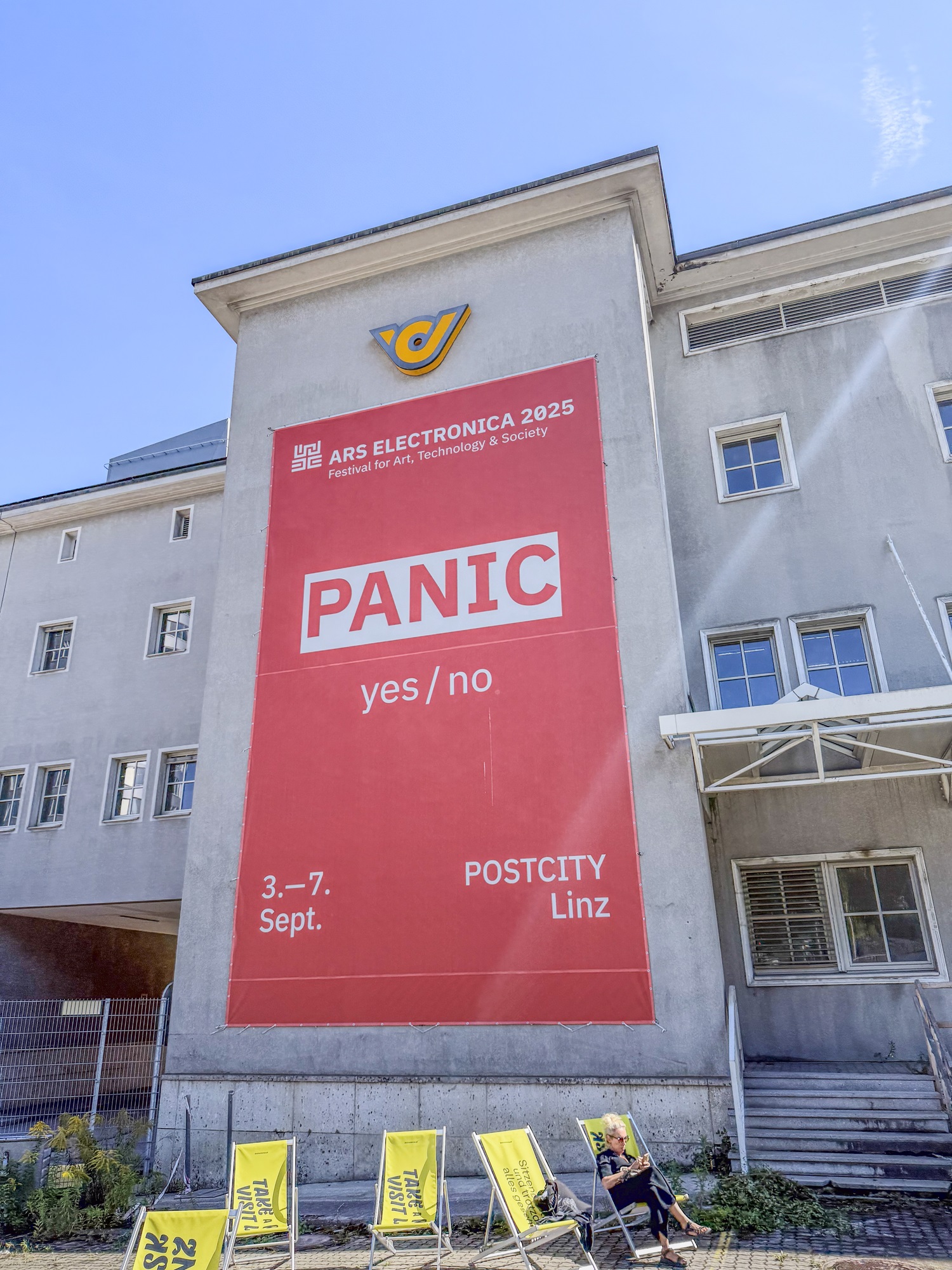 Die Fassade eines Gebäudes ist zu sehen, an der ein großes rotes Banner hängt. Darauf steht „ARS Electronica 2025 – Festival for Art, Technology & Society“ sowie das Motto „PANIC yes / no“. Unten sind die Veranstaltungs-daten „3.–7. Sept.“ und der Ort „POSTCITY Linz“ angegeben. Vor dem Gebäude stehen mehrere gelbe Liegestühle, in einem davon sitzt eine Person.