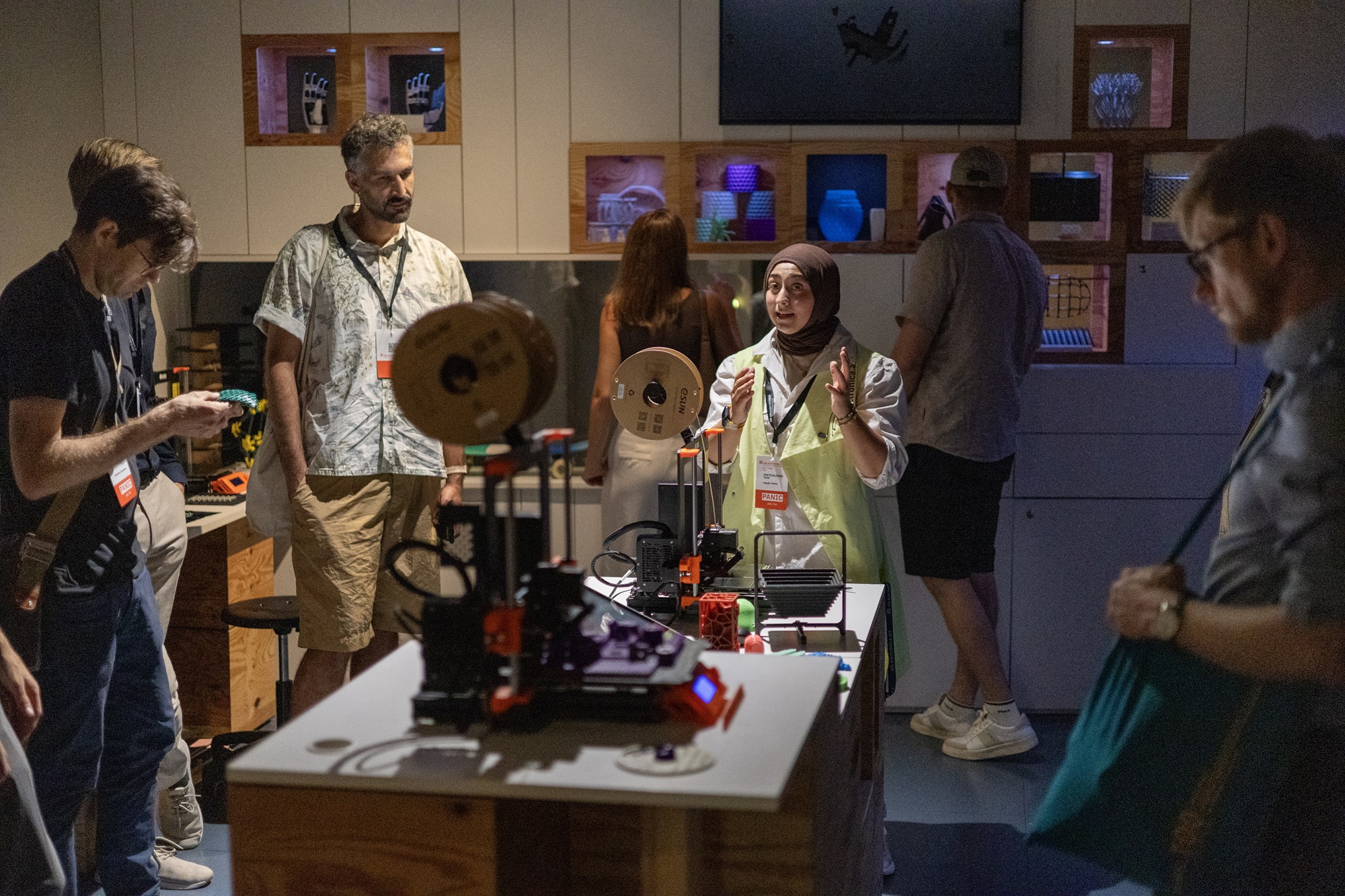 Eine Gruppe von Personen, haben sich um einen Tisch mit mehreren 3D-Druckern versammelt. In der Mitte des Bildes steht eine Frau, die den Besuchern etwas erklärt. Die anderen Personen stehen um den Tisch herum und betrachten die Geräte. Im Hintergrund sind in beleuchteten Regalfächern bunte Objekte ausgestellt.