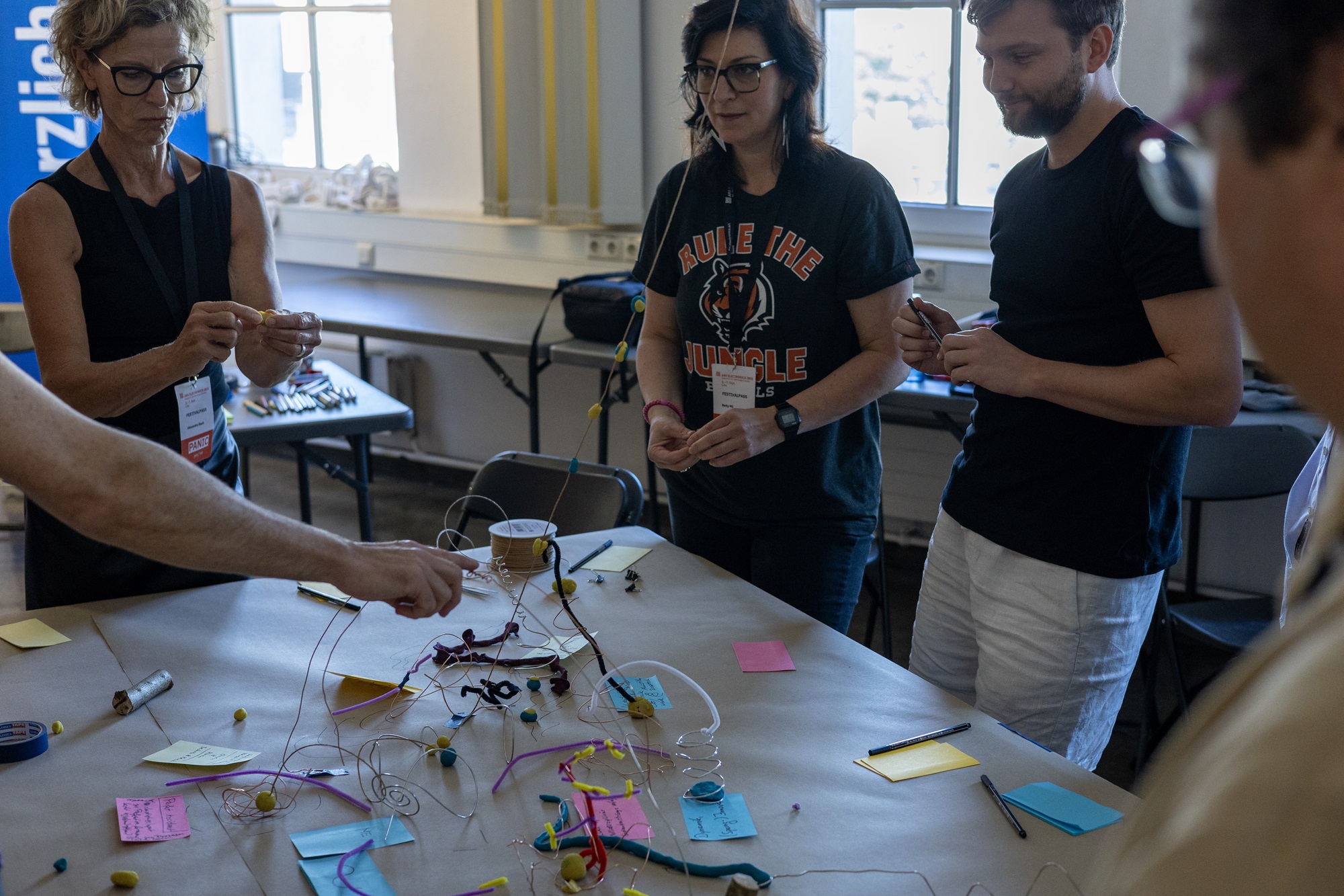 Mehrere Personen stehen um einen Tisch, auf dem bunte Notizzettel, Drähte und Knetstücke liegen. Einige der Teilnehmenden halten diese Materialien in den Händen.
