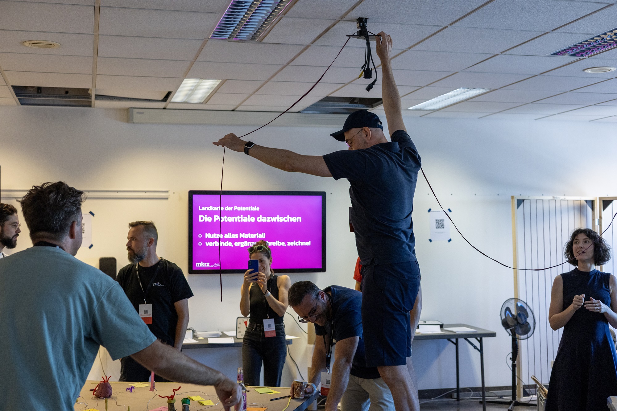 Mehrere Personen sind in einem Raum versammelt. Ein Mann steht auf einem Tisch und befestigt eine Schnur an der Decke, während andere Teilnehmende zuschauen, lachen oder Fotos machen. Im Hintergrund ist ein Bildschirm mit dem Titel „Landkarte der Potentiale“ und Hinweisen zur Materialnutzung zu sehen.
