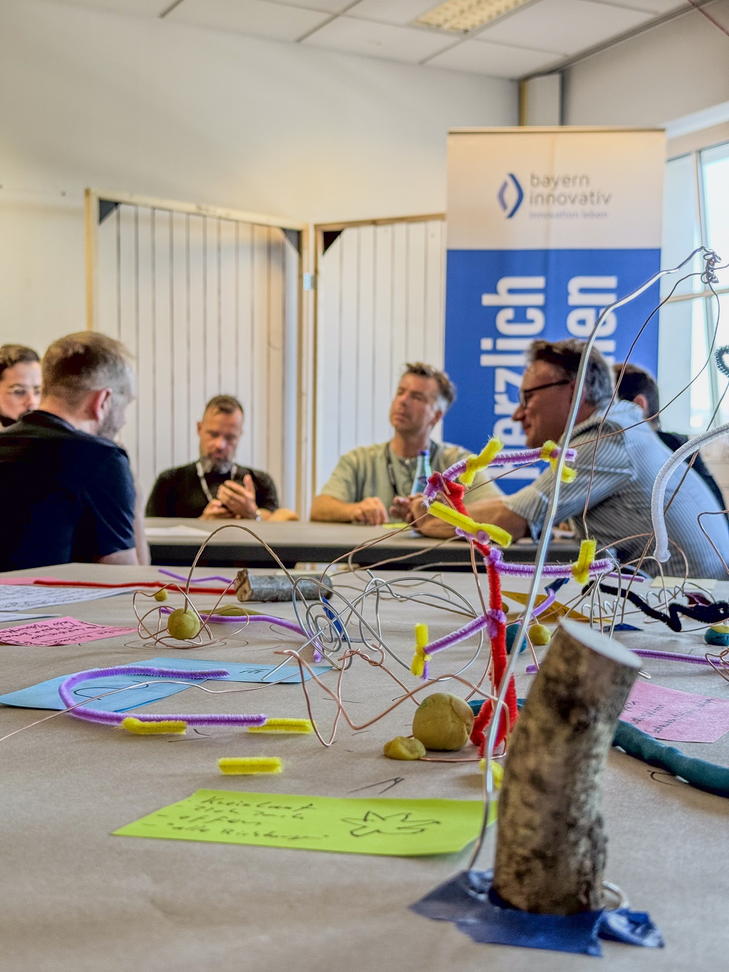 Mehrere Personen sitzen zusammen an einem Tisch und scheinen in ein Gespräch vertieft zu sein. Im Vordergrund sind Bastelutensilien wie Knete zu sehen, die zu kreativen Formen zusammengestellt wurden. Im Hintergrund steht ein Banner mit der Aufschrift „bayern innovativ“.