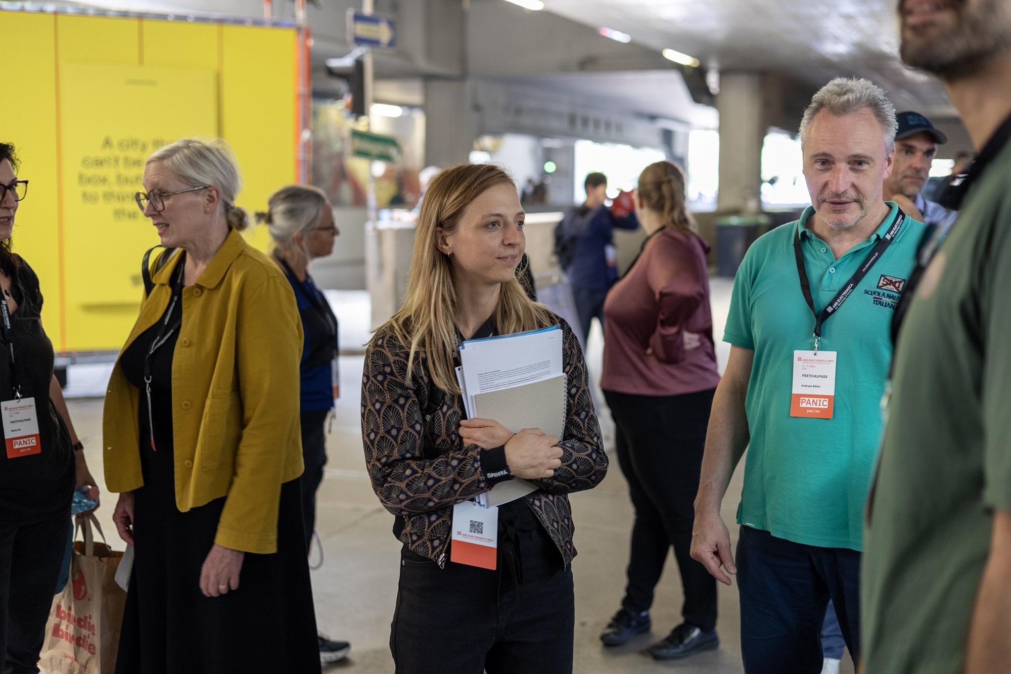 Mehrere Personen stehen in einer Halle zusammen und unterhalten sich. In der Mitte steht Julia Wirnshofer, Projektmanagerin im Innovationsnetzwerk Kreativwirtschaft der Bayern Innovativ GmbH, mit einigen Unterlagen im Arm. Neben ihr steht Andreas Böhm, Innovationsnetzwerk Digitalisierung, Leitung ThinkNet Quantentechnologie, Bayern Innovativ GmbH. Um sie herum sind weitere Menschen im Gespräch.