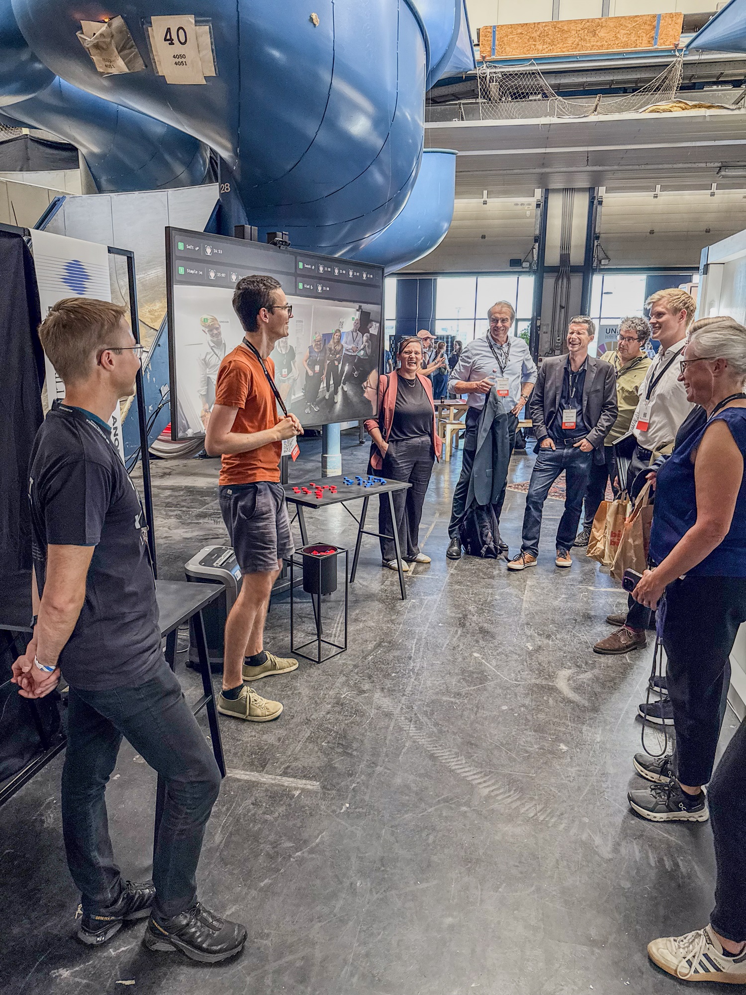 Eine Gruppe von Personen steht in einer Ausstellungshalle um einen Bildschirm, auf dem eine Demonstration gezeigt wird. Zwei Personen vorne erklären etwas, während die anderen zuhören. Im Hintergrund ist eine große blaue Rutsche zu sehen. 