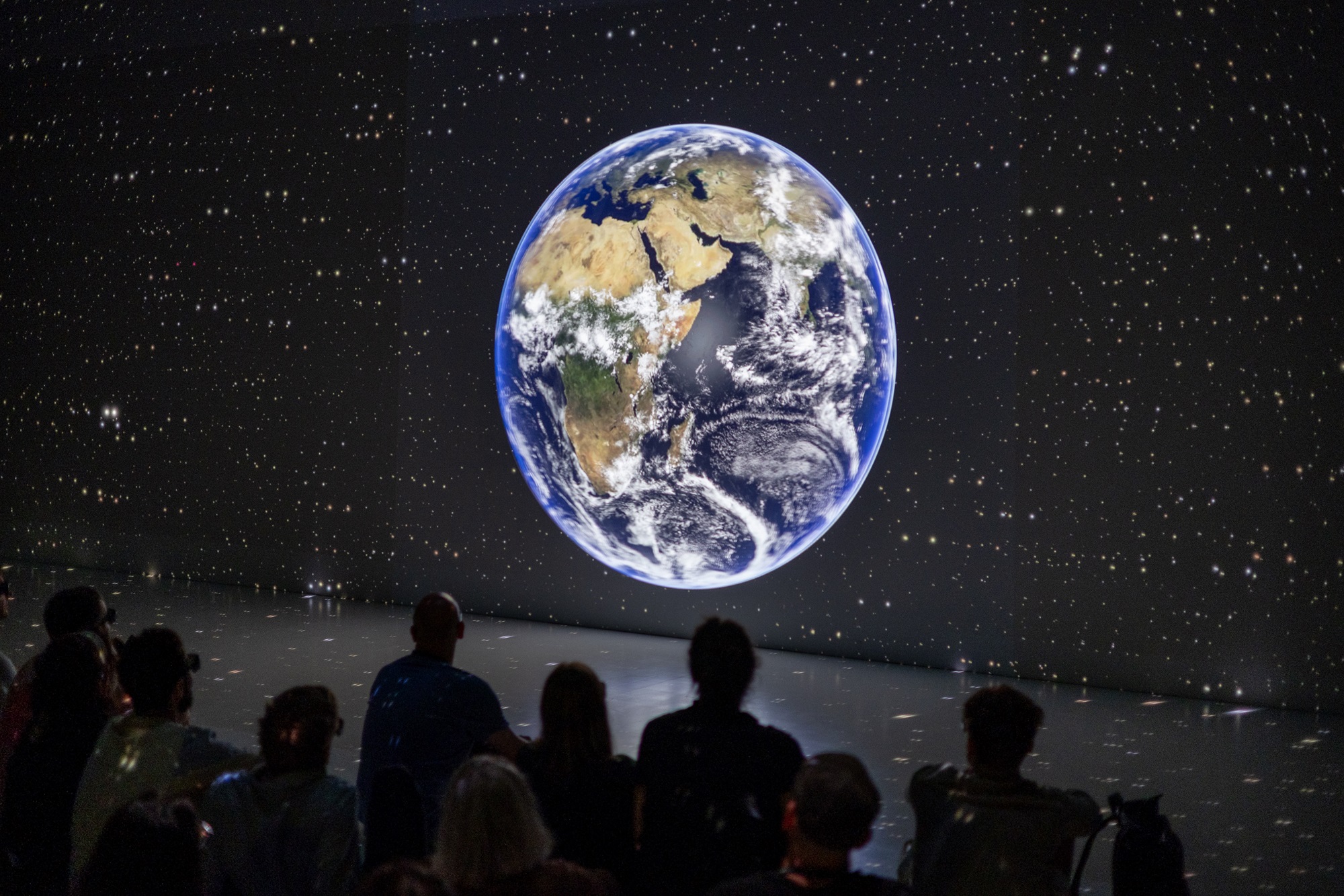 Eine Gruppe von Menschen sitzt nebeneinander in einem abgedunkelten Raum. Sie schauen auf eine riesige Projektion der Erde, die mitten vor einem schwarzen Hintergrund voller Sterne erscheint.