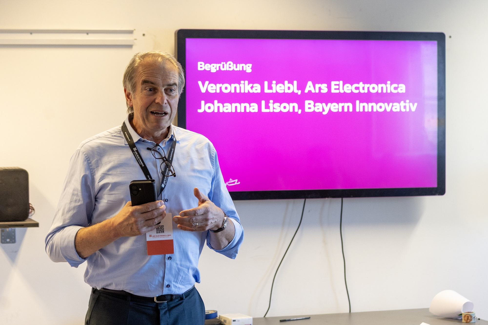 Dr. Markus Eder, Geschäftsführender Vorsitzender, Ministerialdirigent, Leiter der Abteilung Mittelstand, Handwerk, Handel, Freie Berufe im Bayerischen Staatsministerium für Wirtschaft, Landesentwicklung und Energie, steht vor einem Bildschirm und hält einen Vortrag. Im Hintergrund ist auf dem Monitor ein pinkfarbener Begrüßungsbildschirm zu sehen mit den Namen „Veronika Liebl, Ars Electronica“ und „Johanna Lison, Bayern Innovativ“.