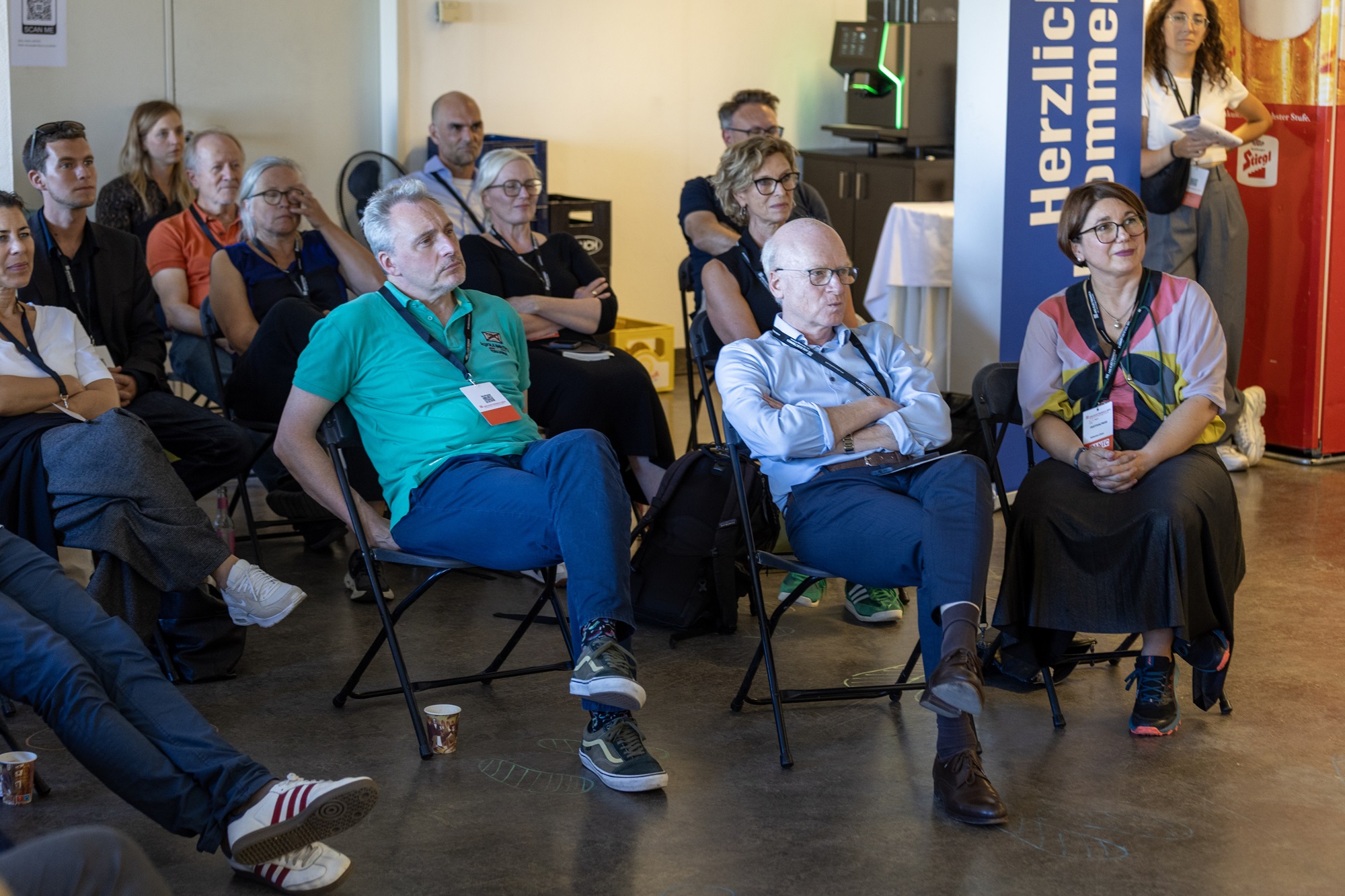 Eine Gruppe von Menschen sitzt in einem Raum und hört aufmerksam zu. Unter ihnen sind Johanna Lison, Projekt- und Innovationsmanagerin im Innovationsnetzwerk Kreativwirtschaft der Bayern Innovativ GmbH, Andreas Böhm, Innovationsnetzwerk Digitalisierung, Leitung ThinkNet Quantentechnologie, Bayern Innovativ GmbH und Matthias Konrad, Mitglied der Geschäftsleitung bei der Bayern Innovativ GmbH. Im Hintergrund ist ein Banner mit der Aufschrift „Herzlich willkommen“ zu sehen.