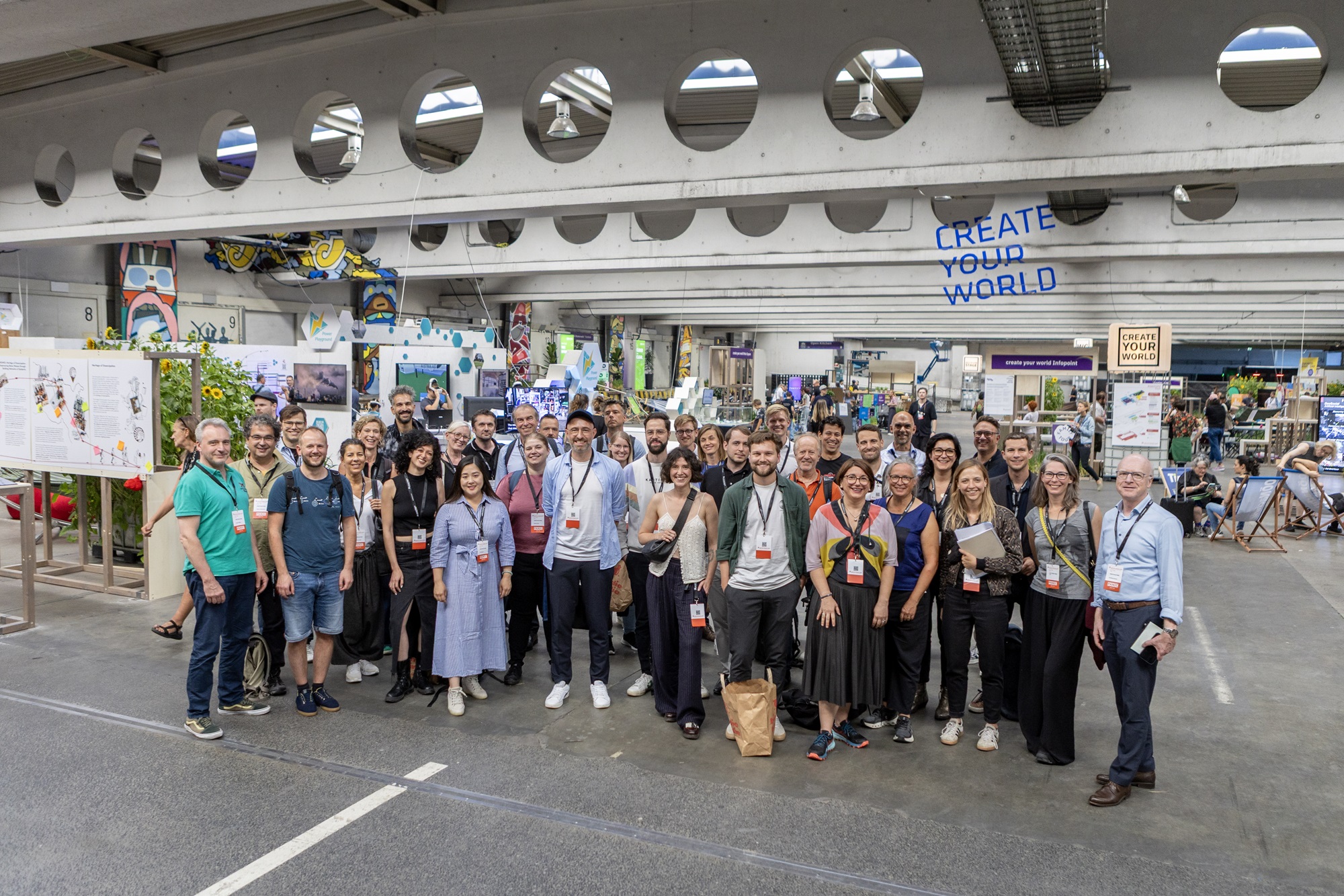 Das Gruppenbild der Teilnehmenden der Cross-Innovation-Reise zur Ars Electronica ist zu sehen. Unter ihnen sind Julia Wirnshofer, Projektmanagerin im Innovationsnetzwerk Kreativwirtschaft der Bayern Innovativ GmbH, Johanna Lison, Projekt- und Innovationsmanagerin im Innovationsnetzwerk Kreativwirtschaft der Bayern Innovativ GmbH, Matthias Konrad, Mitglied der Geschäftsleitung bei der Bayern Innovativ GmbH, sowie Andreas Böhm, Innovationsnetzwerk Digitalisierung, Leitung ThinkNet Quantentechnologie, Bayern Innovativ GmbH. Im Hintergrund sind bunte Stände, Infotafeln und die Schriftzüge „CREATE YOUR WORLD“ zu erkennen.