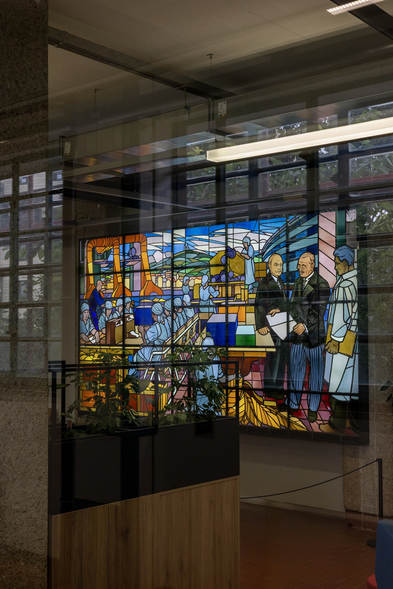 In der Tabakfabrik in Linz ist ein großes, buntes Glasfenster zu sehen, auf dem sich mehrere Personen befinden. Daneben befinden sich klare Fensterflächen, die den Blick nach draußen auf die grünen Bäume freigeben.