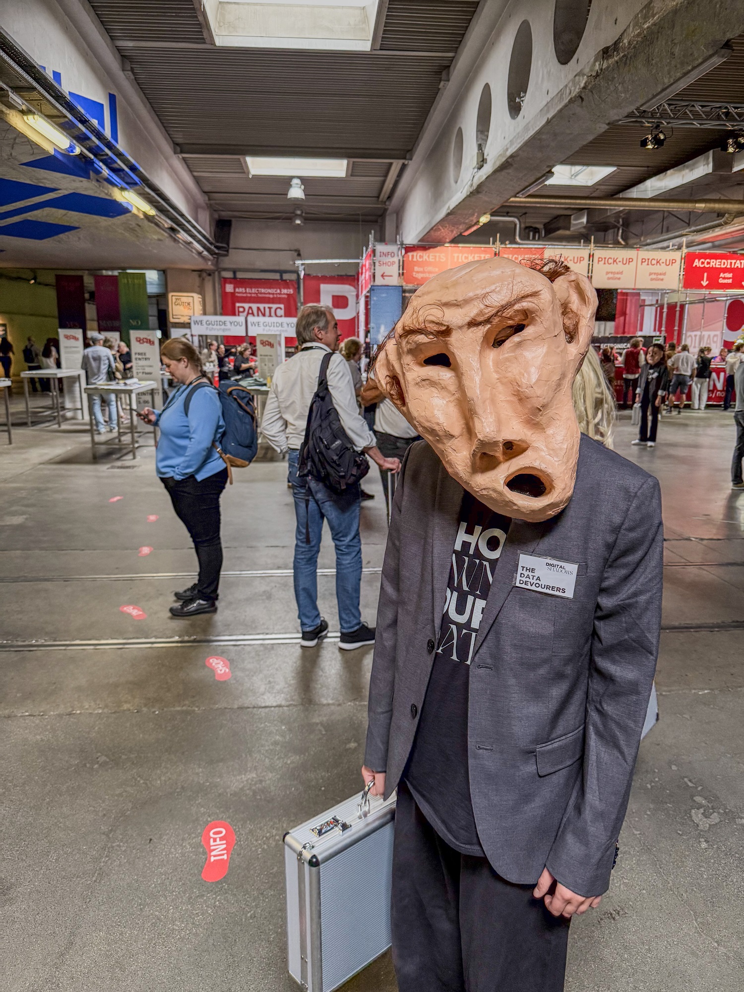 In einer großen Halle steht vorne eine Person mit einer auffälligen Maske, die ein verzerrtes Gesicht darstellt. Sie trägt einen Koffer in der Hand. Im Hintergrund sind mehrere Menschen zu sehen, dazu Schilder für Tickets und Information.