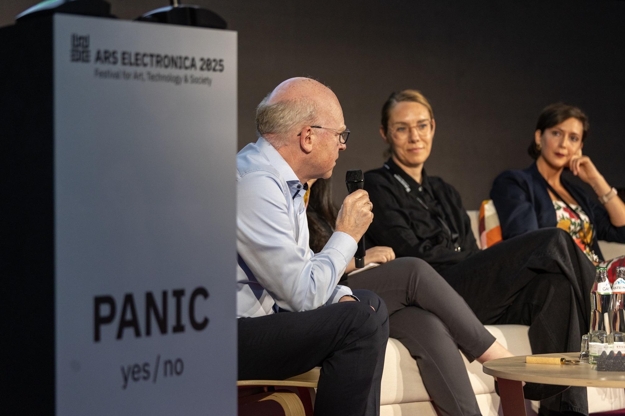 Mehrere Menschen sitzen auf einer Bühne in einer Gesprächsrunde. Matthias Konrad, Mitglied der Geschäftsleitung bei der Bayern Innovativ GmbH, hält ein Mikrofon und spricht, während zwei Frauen neben ihm zuhören. Im Vordergrund steht ein Podium mit der Aufschrift „Ars Electronica 2025“ und dem Motto „Panic yes/no“